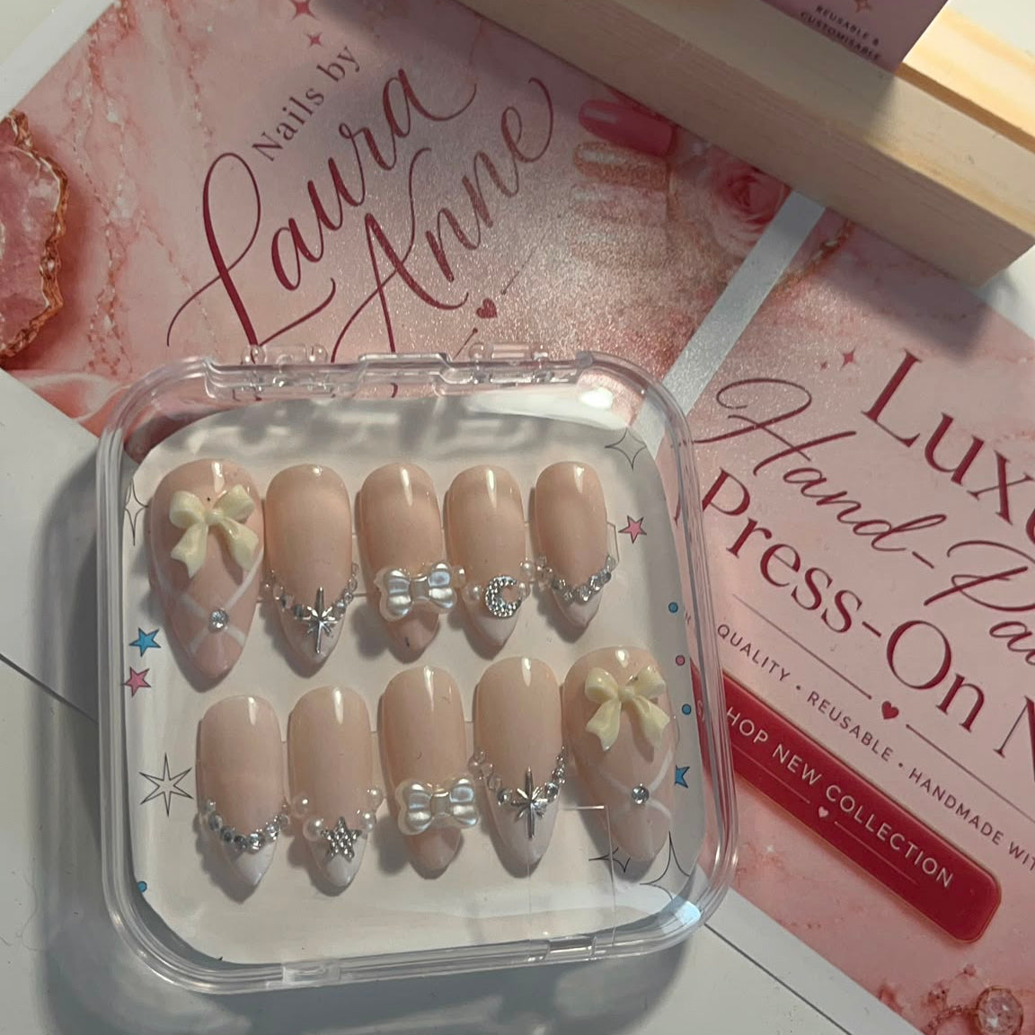 A set of 10 press-on nails in a clear plastic tray, featuring a nude beige base with delicate white floral designs and rhinestone embellishments. The nails are displayed on a white surface alongside pink and white branded packaging cards for 'Laura Anne'.