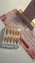 A set of 10 press-on nails in a clear plastic case, displayed on top of pink branded cards and a wooden stand. The nails are a warm nude beige color with small gold leaf designs on two of the tips.