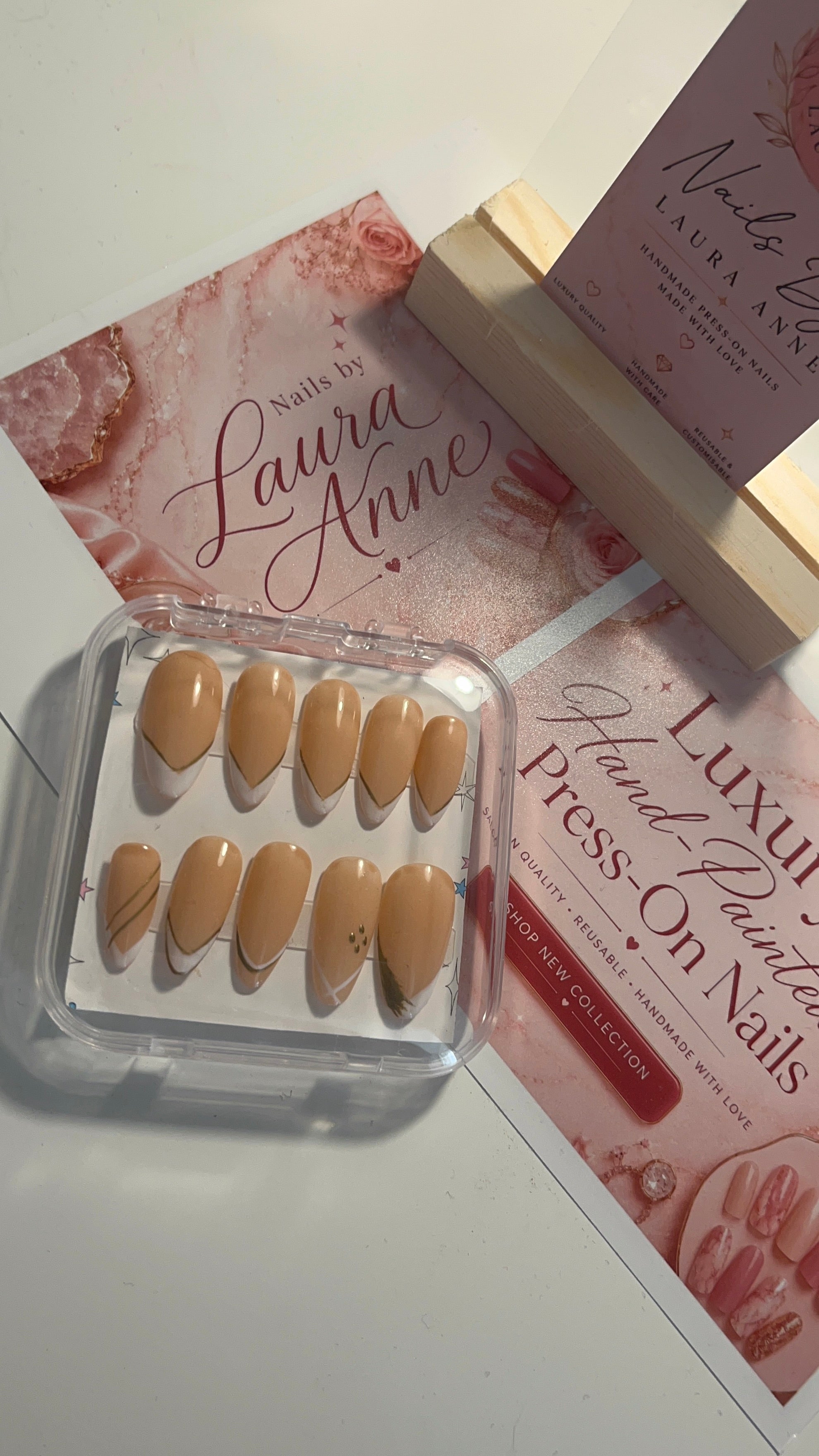 A set of 10 press-on nails in a clear plastic case, displayed on top of pink branded cards and a wooden stand. The nails are a warm nude beige color with small gold leaf designs on two of the tips.