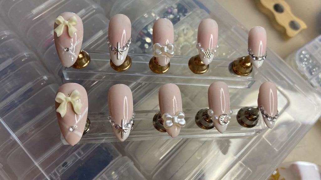 A set of artificial press-on nails displayed on clear plastic stands. The nails have a glossy pale pink base with white French tips, small silver rhinestone accents, pearl details, and two cream-colored bow designs on the lower nails.