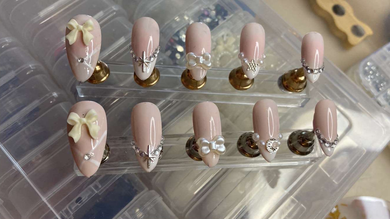 A set of artificial press-on nails displayed on clear plastic stands. The nails have a glossy pale pink base with white French tips, small silver rhinestone accents, pearl details, and two cream-colored bow designs on the lower nails.